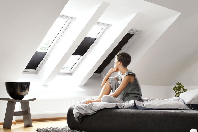 Svetlá podkrovná izba so strešnými oknami a zatemňovacími roletami, žena sedí na posteli