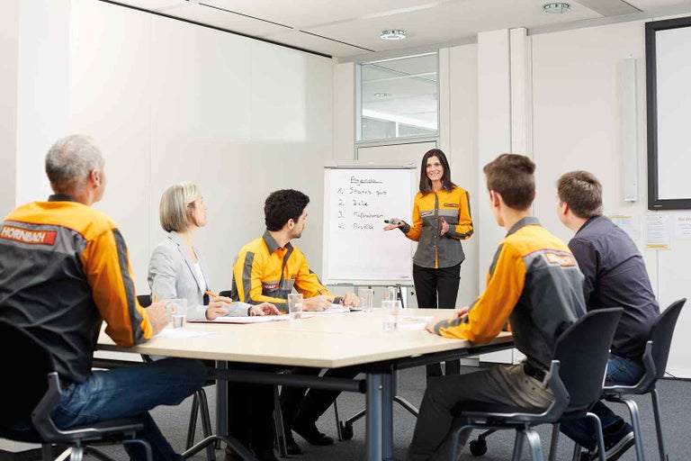 Zasadacia miestnosť s flipchartom a skupinou ľudí v pracovnom oblečení počas prezentácie.