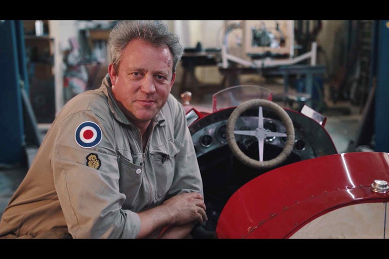 Muž v dielni vedľa červeného veterána závodného auta s dreveným volantom