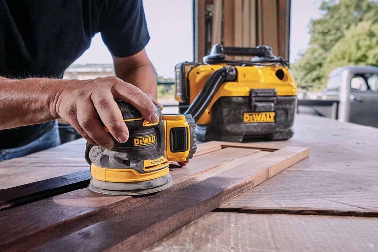 Osoba brúsi drevo excentrickou brúskou značky DeWalt. V pozadí je vysávač.
