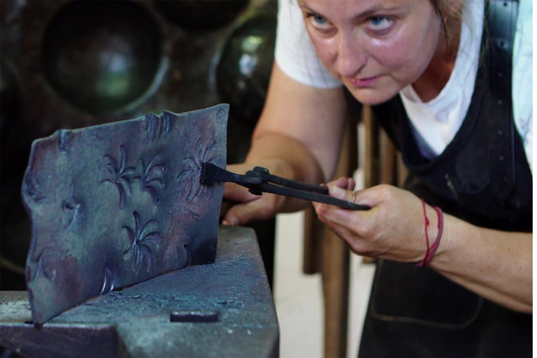 Kovárka spracováva kus kovu s kvetinovým vzorom pomocou klieští na nákovne.