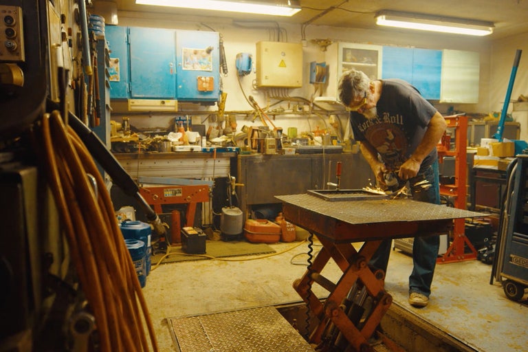Dielňa s mužom, ktorý brúsi kov, pracovný stôl a náradie.