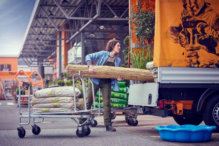 Žena prenáša trstinovú rohož a zeminu pre rastliny s vozíkom k prívesu na parkovisku Hornbach.
