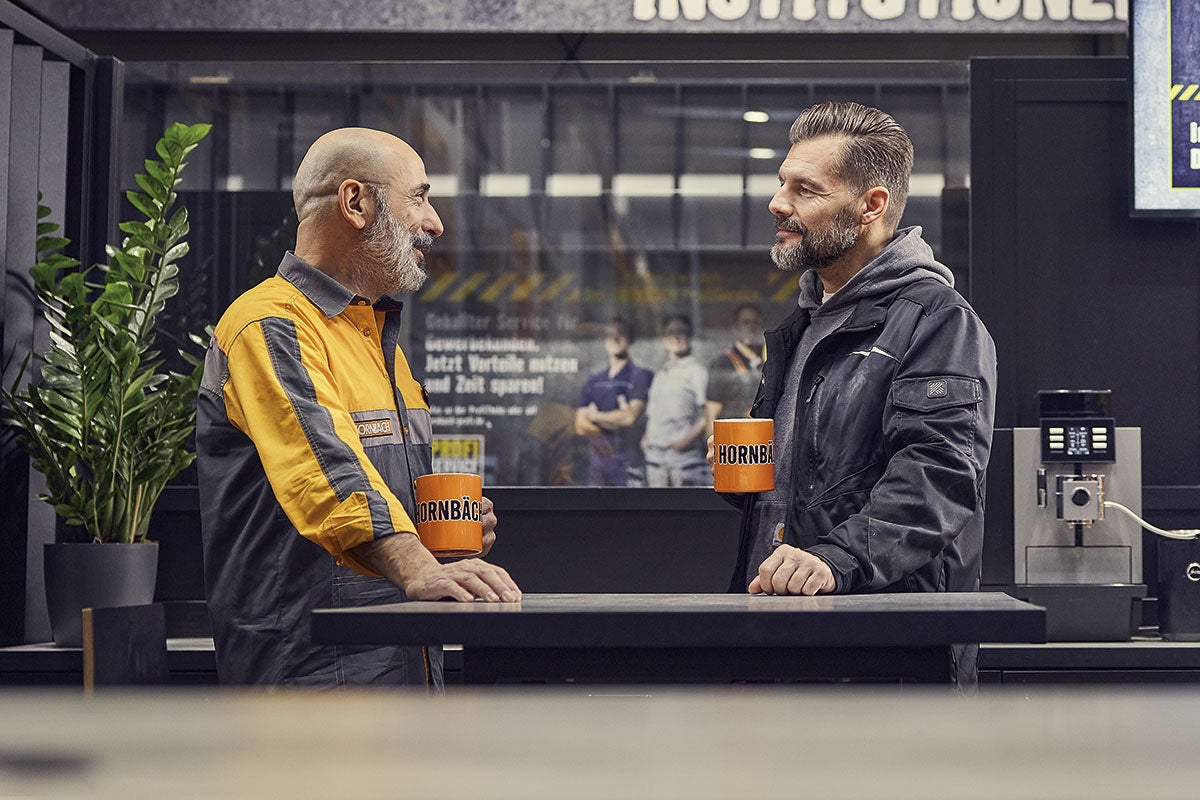 Dvaja muži s hrnčekmi Hornbach sa rozprávajú pri vysokom stole v obchode Hornbach.