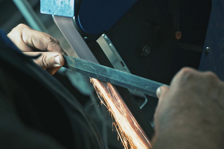 Muž brúsi kov pásovou brúskou, lietajú iskry.