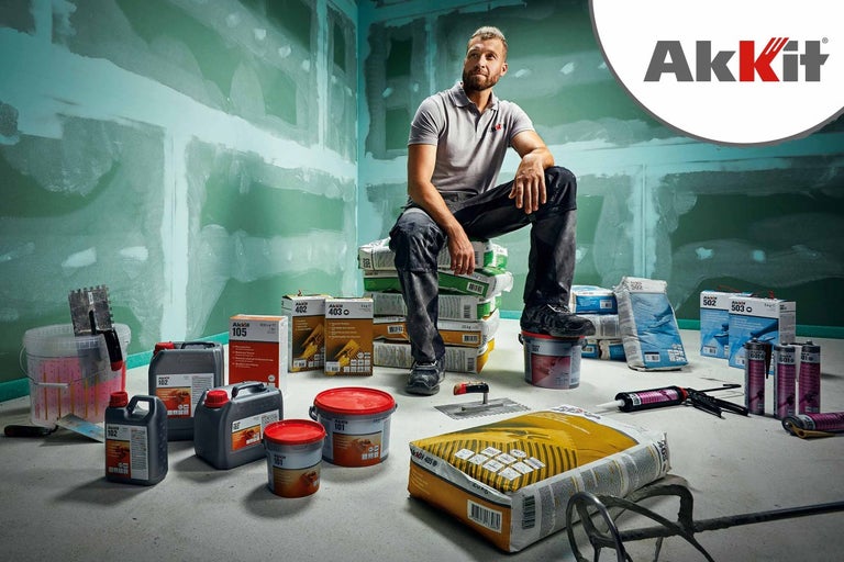 Muž sedí na vreciach s produktmi Akkit v miestnosti s omietnutými stenami, obklopený rôznymi produktmi Akkit, ako sú lepidlá a tesniace hmoty