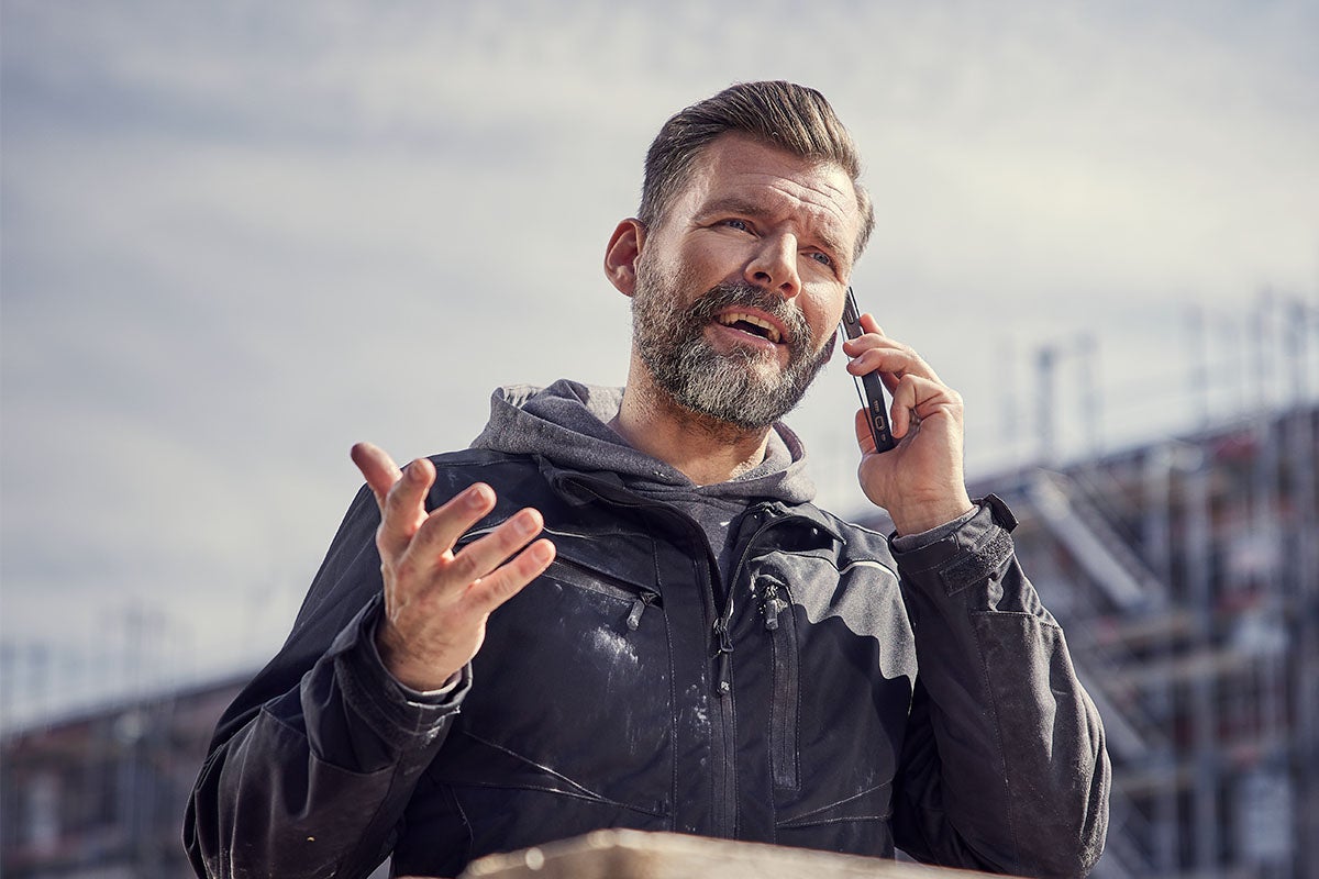 Bradatý muž v tmavej pracovnej bunde a mikine telefonuje na stavenisku so smartfónom a pritom gestikuluje.