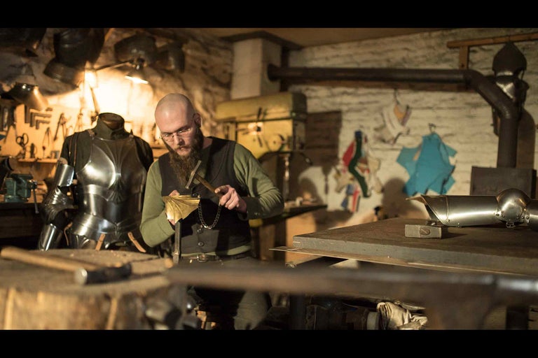 Dielňa s kováčom, ktorý spracováva kov, náradie a brnenie v pozadí