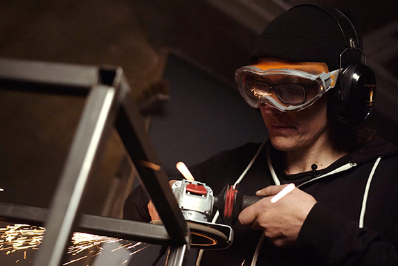 Osoba brúsi kov pomocou brúsky, pričom má ochranné okuliare a chrániče sluchu.