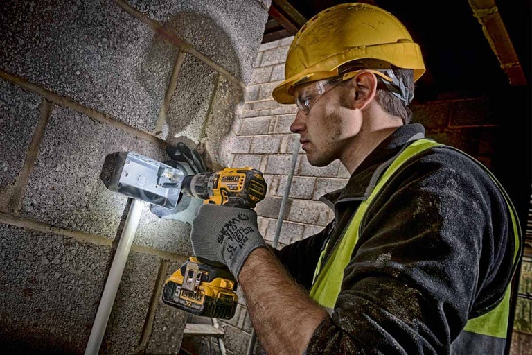 Remeselník so žltou helmou a ochrannými okuliarmi vŕta akumulátorovým vŕtacím skrutkovačom otvor do steny pre odbočnú krabicu.
