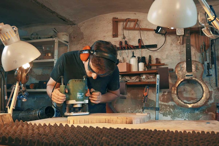 Muž pracuje s hornou frézou na kuse dreva v dielni. Má ochranné okuliare a chrániče sluchu.