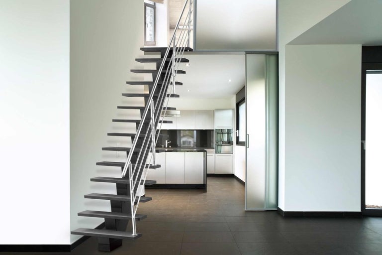 Interiérová snímka moderného domu so samonosným dreveným schodiskom a nerezovým zábradlím, ktoré vedie do kuchyne.