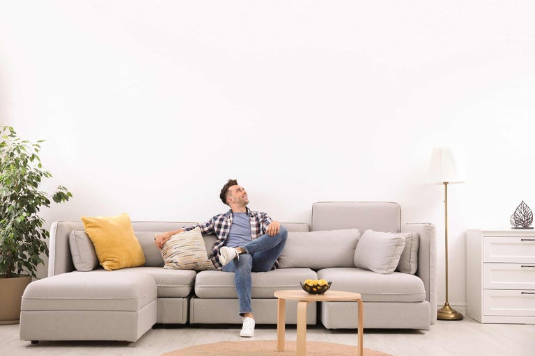 Muž sedí uvoľnene na sivej rohovej pohovke v jasnej obývačke.