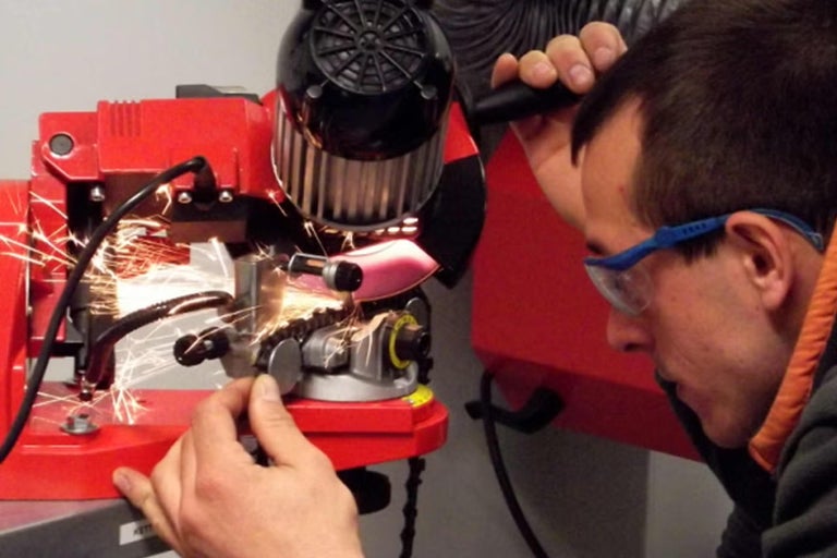 Muž v ochranných okuliaroch brúsi pílovú reťaz na elektrickej brúske na reťaze, pričom odlietavajú iskry.