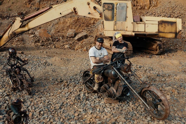 Scéna s bagrom, mužom a ženou a dvoma motocyklami.