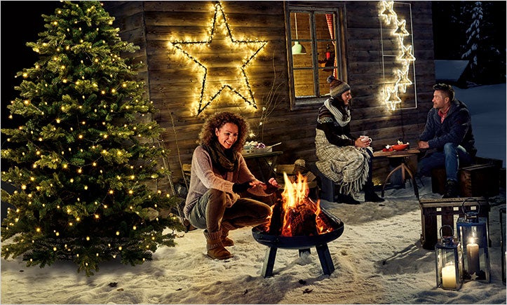 Zimná scéna s ľuďmi pri táboráku, vianočný stromček so svetlami, hviezdy ako dekorácia a drevený dom