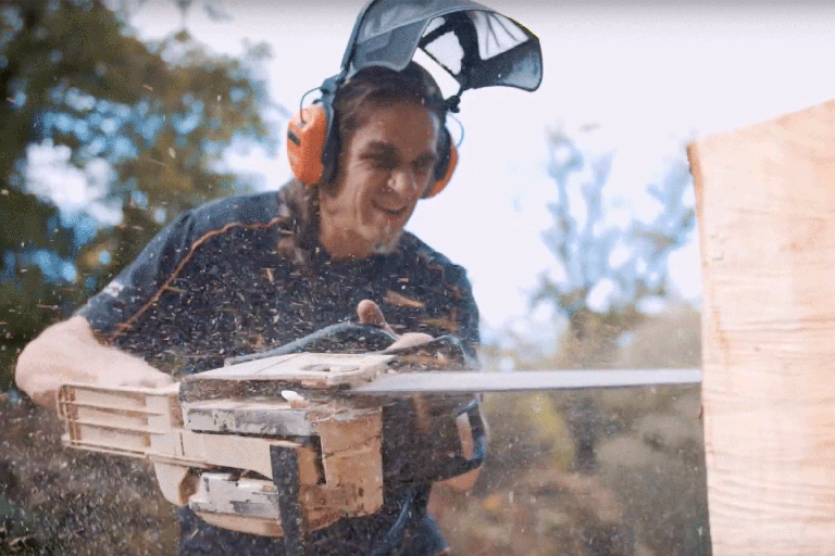 Muž pília drevo motorovou pílou a má na sebe chrániče sluchu a tvárový štít.