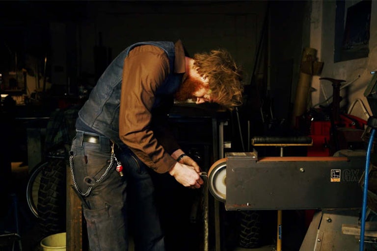 Muž brúsi kovový kus na pásovej brúske v dielni