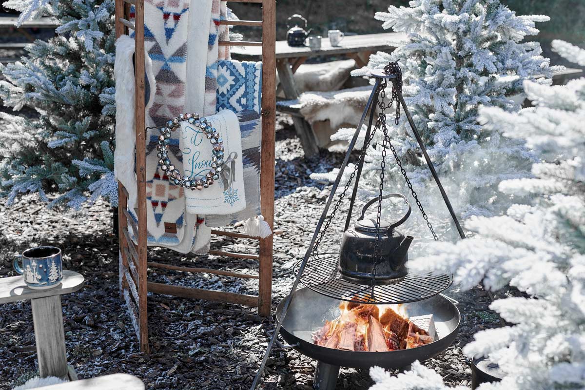 Zimná záhradná scéna s ohniskom, kotlíkom, prikrývkami na rebríku a zasneženými stromami.