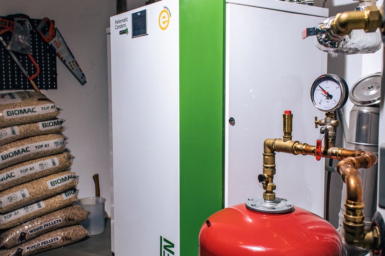 Peletové kúrenie vedľa vriec s drevenými peletami, tlakomeru a náradia na stene.