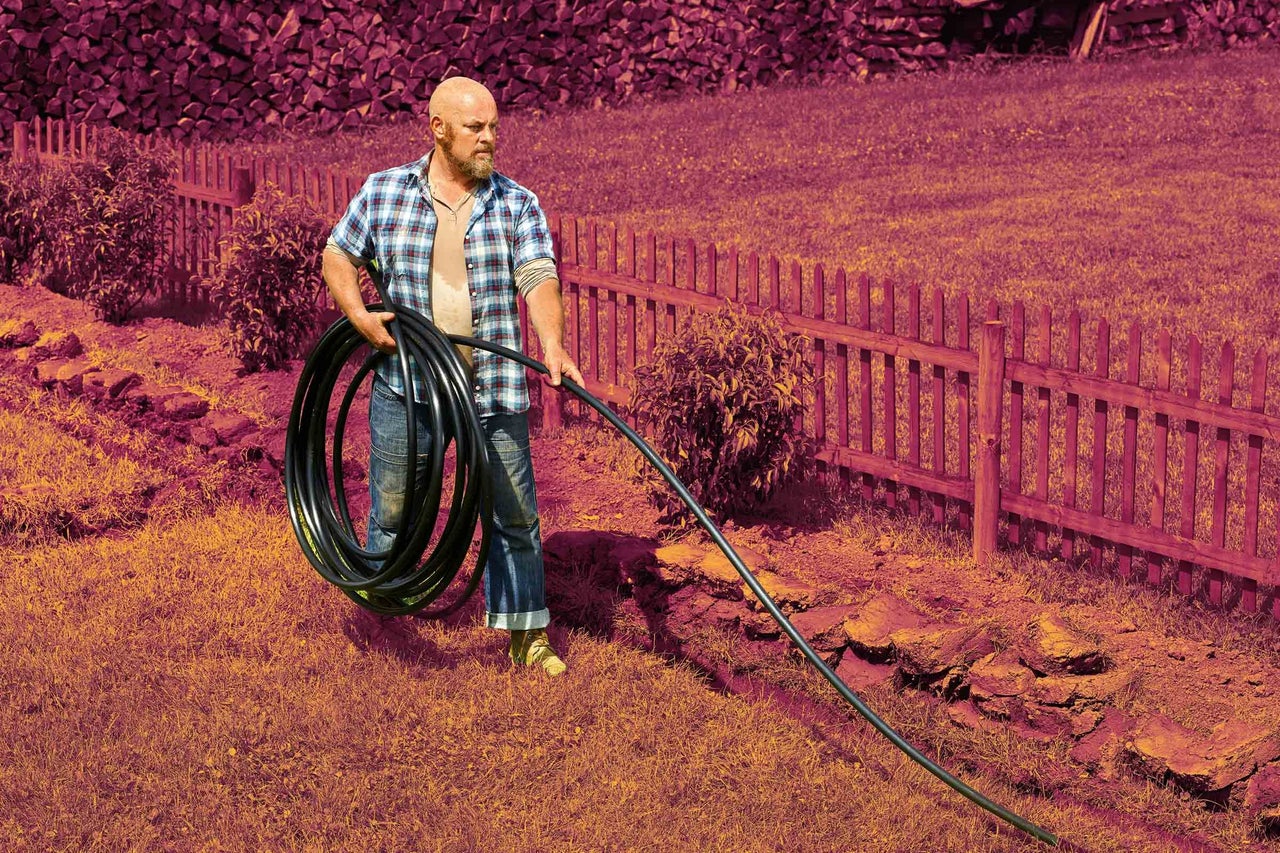 Muž nesie rolu čiernej polyetylénovej rúry na zavlažovanie v záhrade s dreveným plotom.