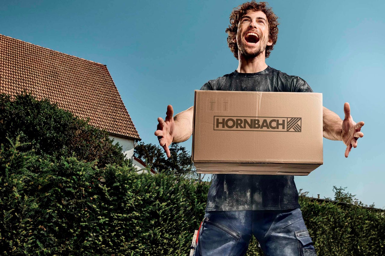 Muž drží krabicu s logom Hornbach na pozadí domu a modrej oblohy.
