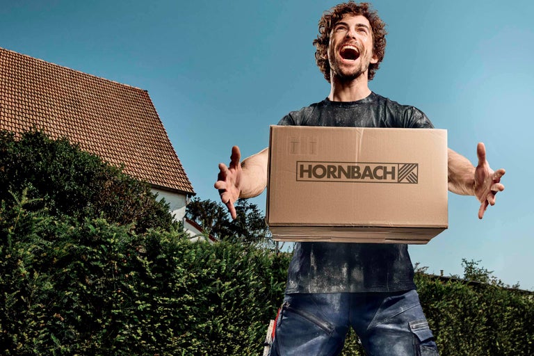 Muž drží krabicu s logom Hornbach na pozadí domu a modrej oblohy.