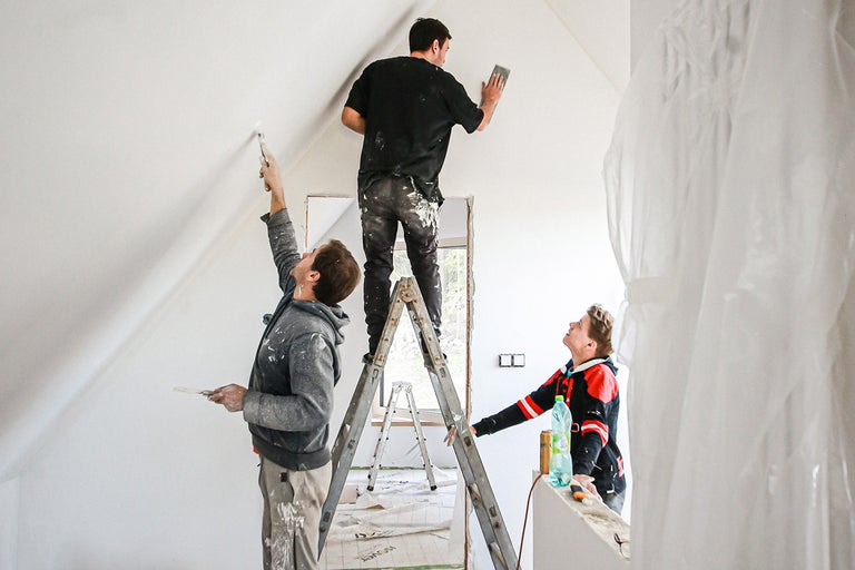 Traja remeselníci renovujú izbu pomocou rebríkov, farby a brúsnych nástrojov.