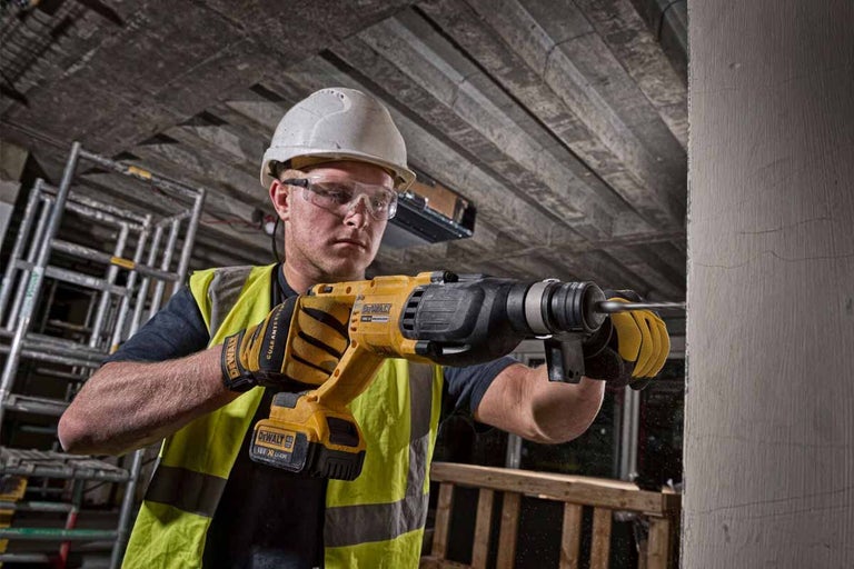 Muž vŕta vŕtačkou otvor do steny. Má na sebe ochrannú helmu, ochranné okuliare a ochranné rukavice.