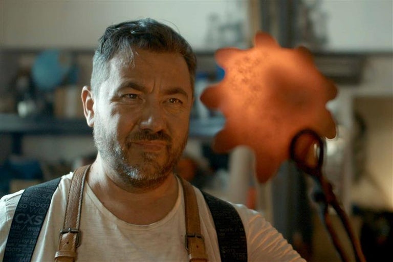 Muž so zásterou drží horúci obrobok kliešťami.