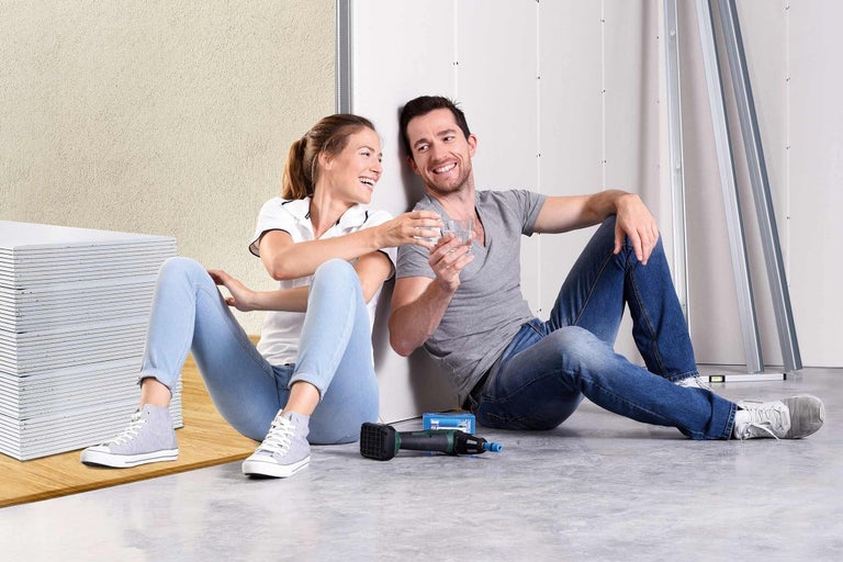 Pár sedí uvoľnene vedľa sadrokartónových dosiek, kovových profilov a akumulátorového skrutkovača na sadrokartón