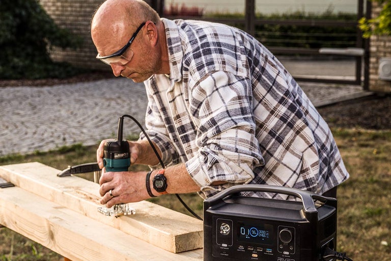 Muž používa hornú frézku na drevenej doske vonku, s prenosnou napájacou stanicou v popredí.
