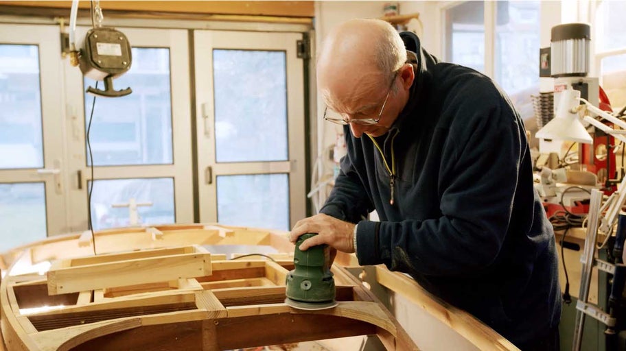 Muž brúsi drevenú loď elektrickou brúskou v dielni.