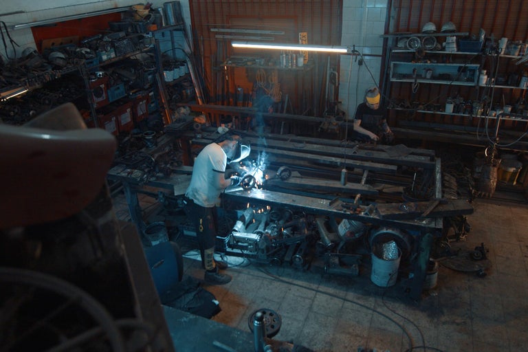 Dielňa s dvoma remeselníkmi, ktorí zvárajú a pracujú na pracovných stoloch, regály s náradím a materiálom.