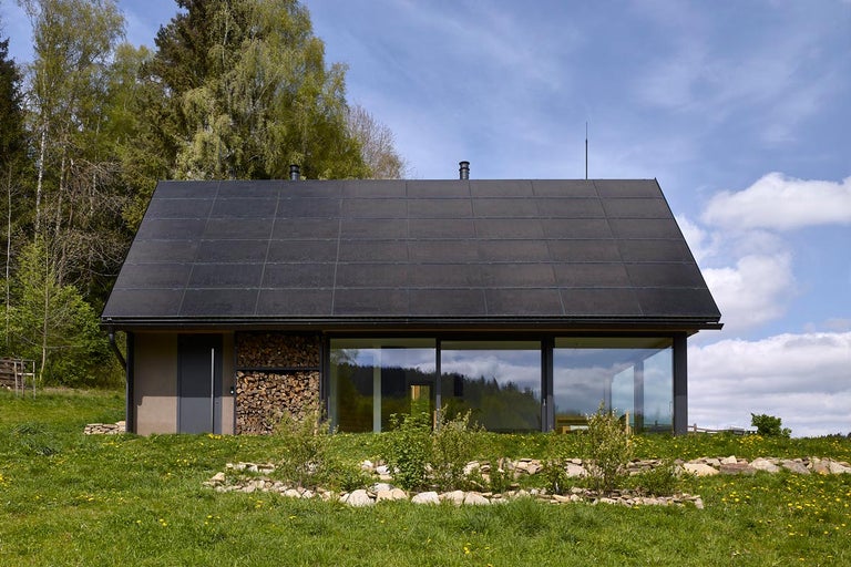 Moderný dom so solárnymi panelmi na streche, skladom dreva a veľkými oknami.