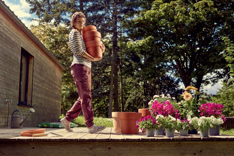 Žena nesie hromadu kvetináčov na drevenej terase s kvetinami a záhradnými doplnkami.