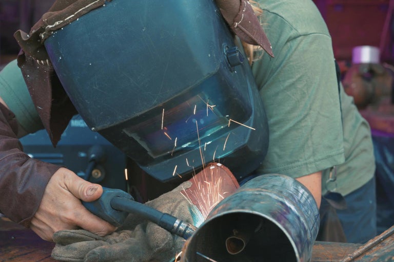 Osoba zvára kovovú rúrku s ochrannou maskou a rukavicami.