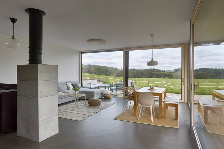 Svetlá obývacia izba s krbom, pohovkou a jedálenským stolom pred veľkým oknom s výhľadom do prírody.