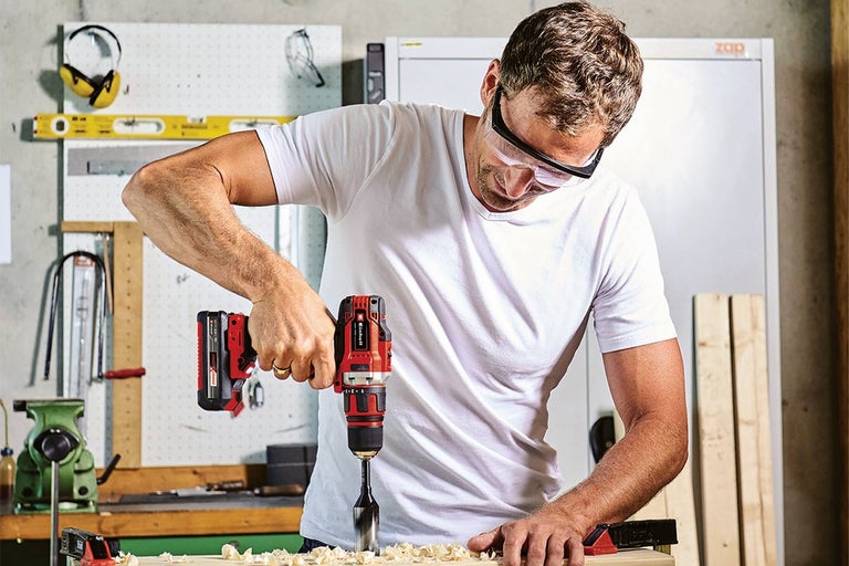 Muž vŕta akumulátorovou vŕtačkou s nadstavcom otvor do kusa dreva. Má ochranné okuliare.