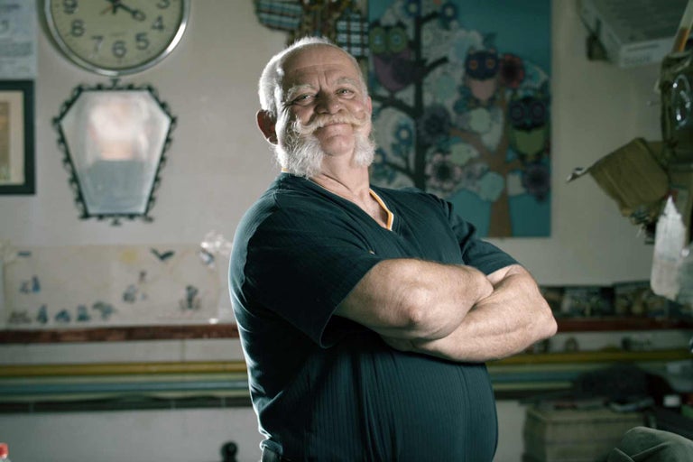 Portrét muža s bradou a prekríženými rukami v miestnosti s hodinami, zrkadlom a obrazom.