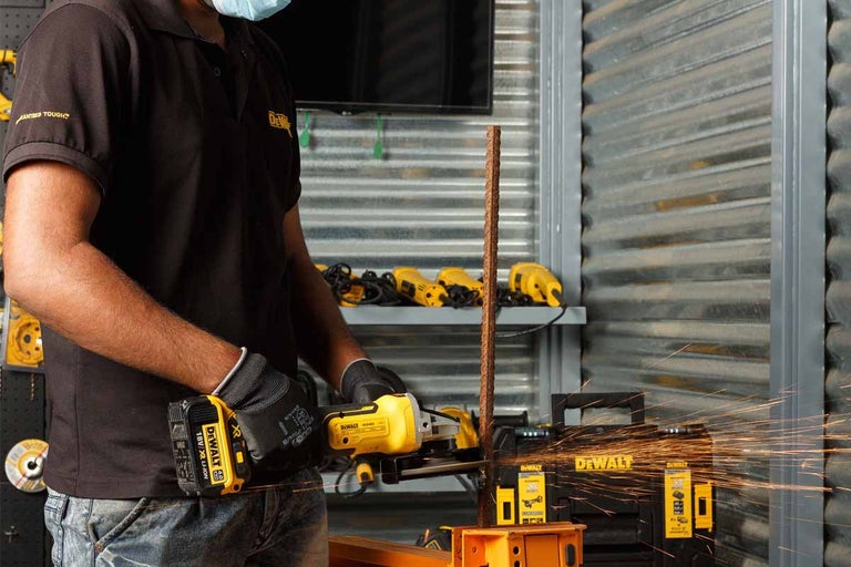 Muž reže uhlovou brúskou kus kovu, pričom lietajú iskry. V pozadí je náradie a logo DeWalt.