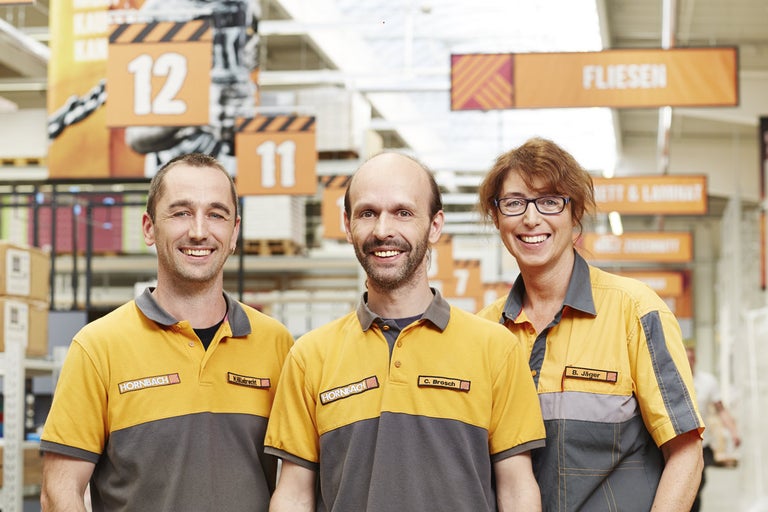 Traja zamestnanci Hornbach v pracovnom odeve pred regálmi so stavebným materiálom.