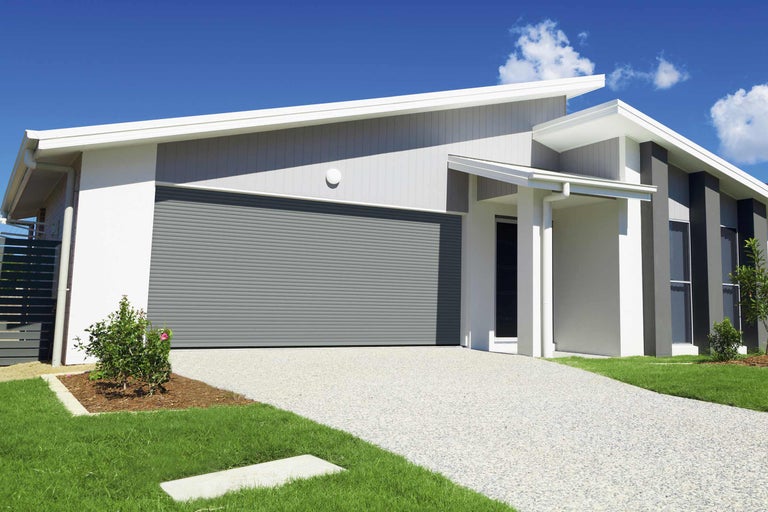 Moderný dom s garážou so sivou rolovacou bránou, dláždenou cestou a udržiavanou záhradou.