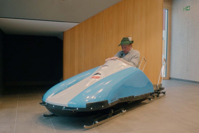 Muž sedí v modro-bielej pretekárskej sánke v interiéri