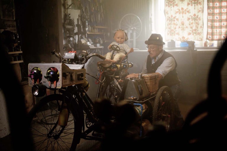 Muž s klobúkom a okuliarmi stojí vedľa upraveného bicykla s košíkom a bábikou ako vodičom v dielni.