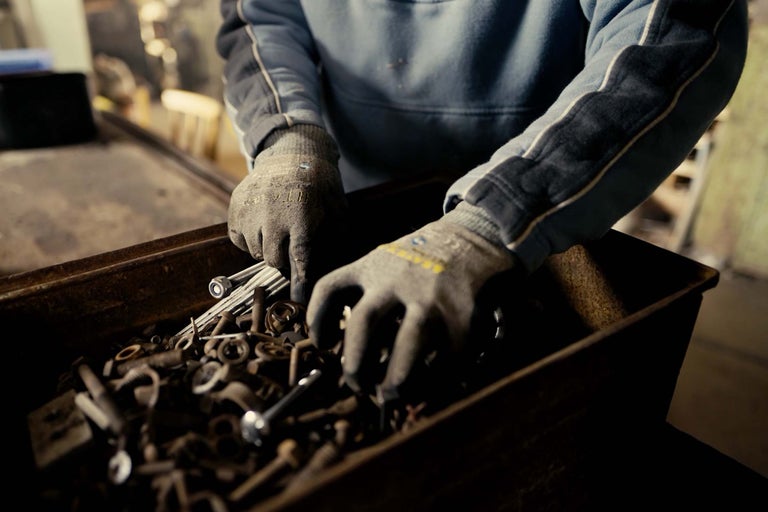 Osoba prehľadáva debnu so skrutkami a maticami v pracovných rukaviciach.