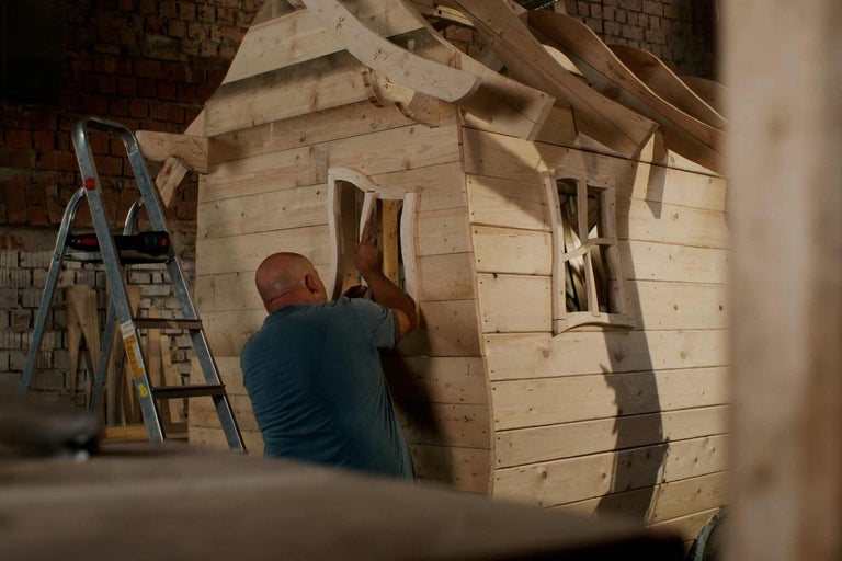 Drevený domček pre deti stavia muž. Vedľa stojí rebrík.