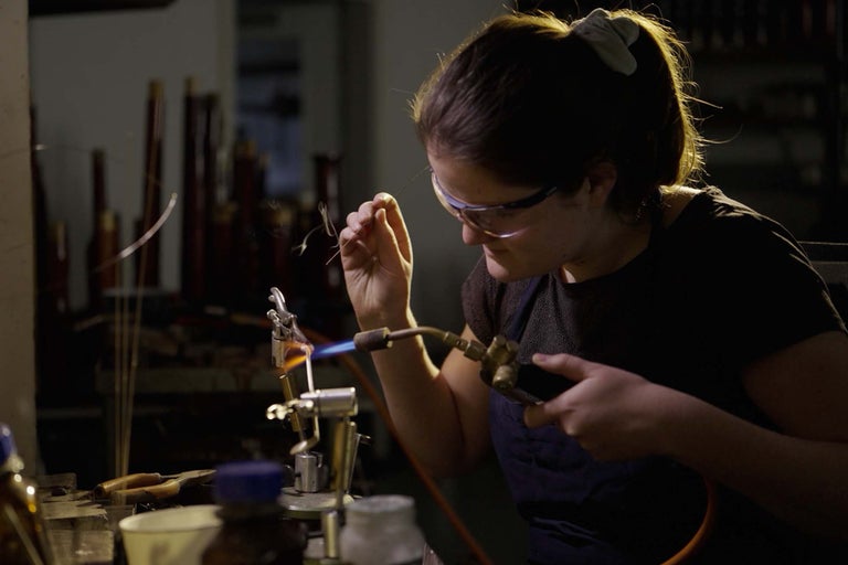 Žena s ochrannými okuliarmi a spájkovacou lampou pri spracovaní skla.