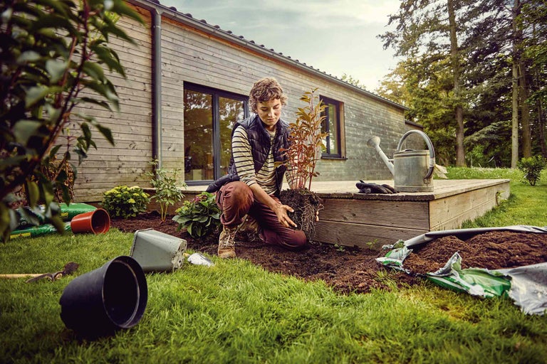 Osoba sadí rastlinu v záhrade, s kvetináčmi, hlinou a kanvou v blízkosti.