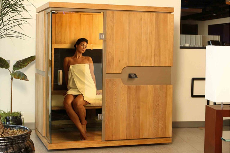 Drevená sauna so sklenenými dverami a sediaca žena s uterákom